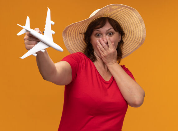 中间身穿红色t恤 头戴夏帽的中年妇女 展示玩具飞机惊讶地捂着嘴 手站在橙色的墙上封面帽子站