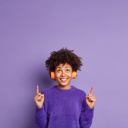 产品休闲套头衫中快乐的非裔美国女人的垂直镜头表明上面的文字空间上有正面的表情微笑着高兴地戴上立体声耳机非洲积极手势
