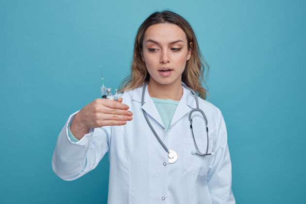年轻印象深刻的年轻女医生穿着医用长袍 戴着听诊器 围着脖子拿着针管 看着安瓿和疫苗瓶注射器长袍穿着