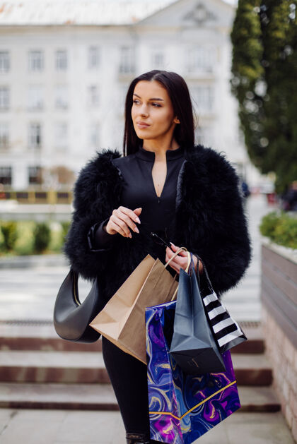 顾客一个棕色头发的女人穿着黑色的衣服 手里拿着五颜六色的有图案的购物袋 在一次成功的购物狂欢中走在外面 她享受着一天的温暖女士服装城市