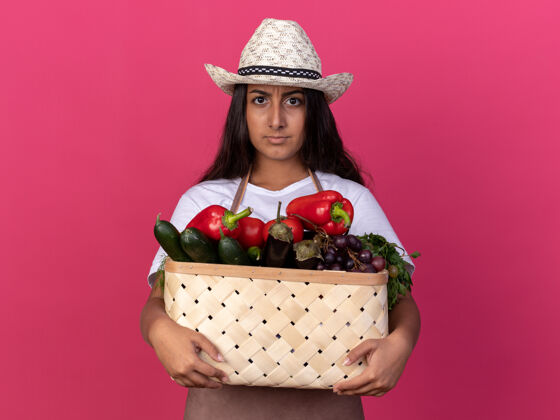 立场穿着围裙 戴着夏帽的年轻园丁女孩站在粉色的墙上 手里拿着装满蔬菜的箱子 严肃的脸充分帽子花园