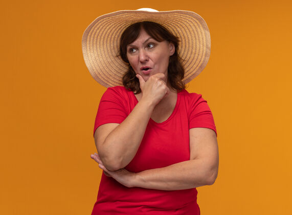 沉思身穿红色t恤 头戴夏帽的中年妇女站在橙色的墙上 一边沉思一边思考帽子女人中年