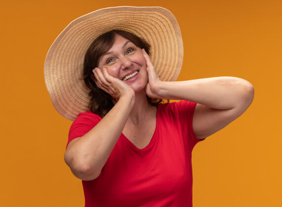 年龄一位身穿红色t恤 头戴夏帽的中年妇女站在橙色的墙上 脸上挂着幸福的表情 两手放在脸颊上站脸颊女人