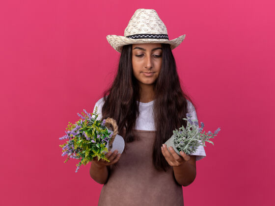 年轻穿着围裙 戴着夏帽的年轻园丁女孩 手里拿着盆栽植物 站在粉红色的墙上严肃地看着它们围裙严肃