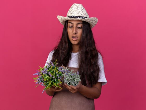 花园年轻的园丁女孩围着围裙 戴着夏帽 手里拿着盆栽植物 站在粉色的墙上惊讶地看着它们惊喜年轻的锅