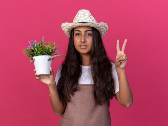 表演穿着围裙 戴着夏帽 手持盆栽的年轻园丁女孩微笑着 粉红色的墙上立着v字标志围裙花园花盆