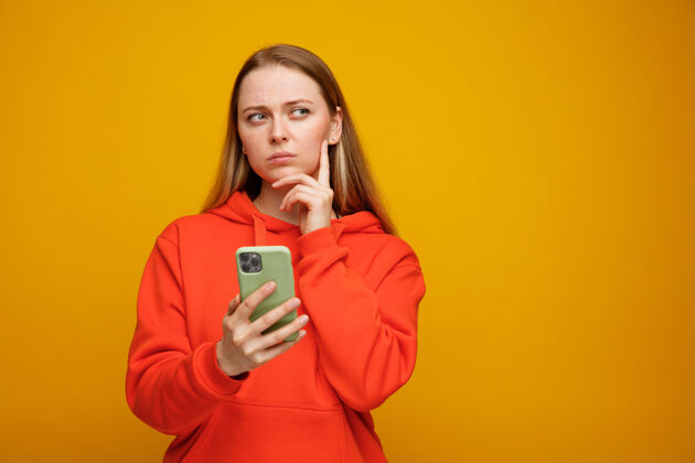 橙色体贴的年轻金发女人 手放在下巴上 手拿着手机 看着手机拿着电话手机