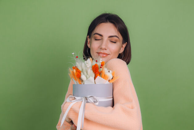 圆形年轻女子穿着休闲桃色毛衣隔离在绿橄榄墙上手持橙白色花盒组成的棉花 吉普赛拉小麦和拉古鲁斯作为礼物开心惊喜积极欢乐美丽