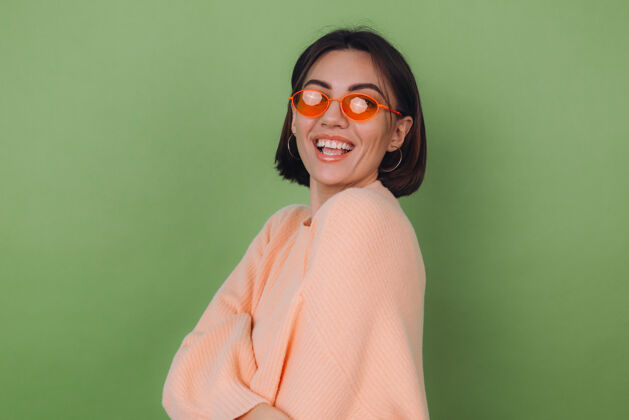 快乐年轻时尚的女士穿着休闲的桃色毛衣 戴着橙色的眼镜 隔着绿橄榄色的墙壁 开心的 积极的笑着 围着复制空间转时尚爱情表情