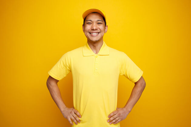 帽子快乐的年轻送货员戴着帽子 穿着制服 手放在腰上快乐穿黄色
