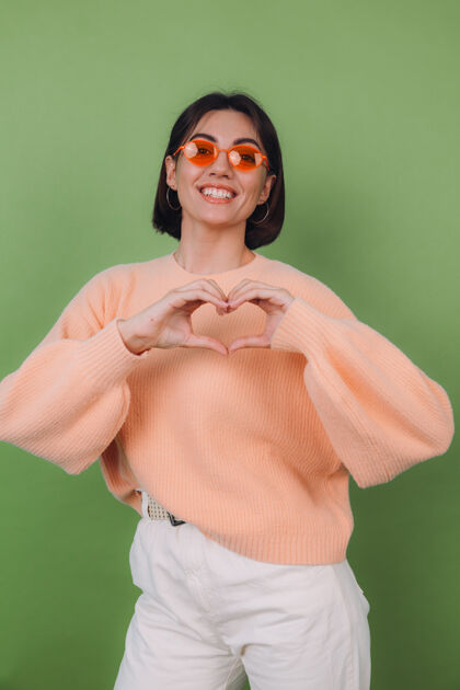 心年轻女子穿着休闲桃色毛衣 戴着橘色眼镜 隔离在绿橄榄色的墙上 展示着用心用手爱的概念复制空间情人手势形状