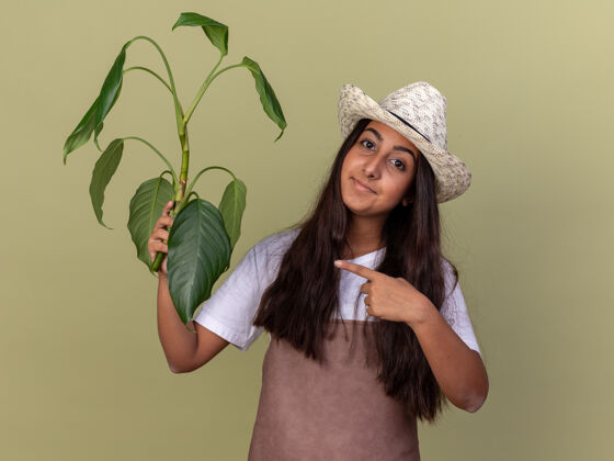 目录穿着围裙 戴着夏帽的年轻园丁女孩拿着植物 用食指指着它 微笑着自信地站在绿色的墙上围裙帽子手指