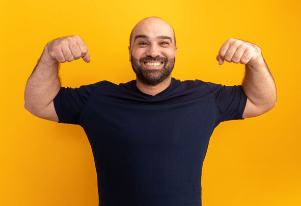胡须一个留着胡子的穿着海军t恤的男人举起拳头 像一个胜利者 站在橘色的墙上 开心而兴奋地微笑着宽阔站着兴奋