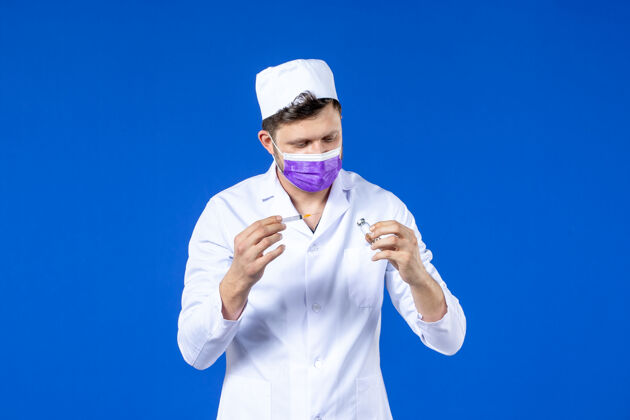 药品前视图中的男医生穿着医疗服和口罩持有疫苗和注射在蓝色病毒疫苗颜色