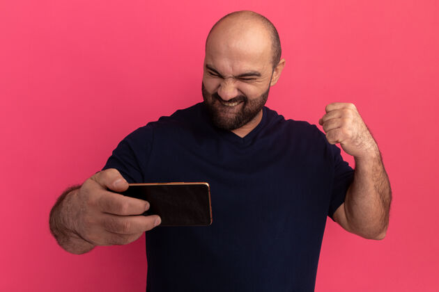 握紧一个留着胡子的男人穿着海军蓝t恤 拿着智能手机看着它 高兴而兴奋地握紧拳头站在粉色的墙上拳头移动胡须