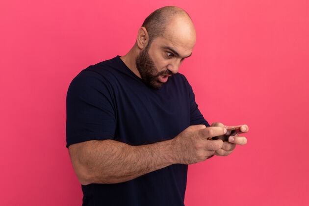 移动一个留着胡子的男人穿着海军蓝t恤 用智能手机玩游戏 站在粉色的墙上 看上去既惊讶又困惑惊喜玩游戏