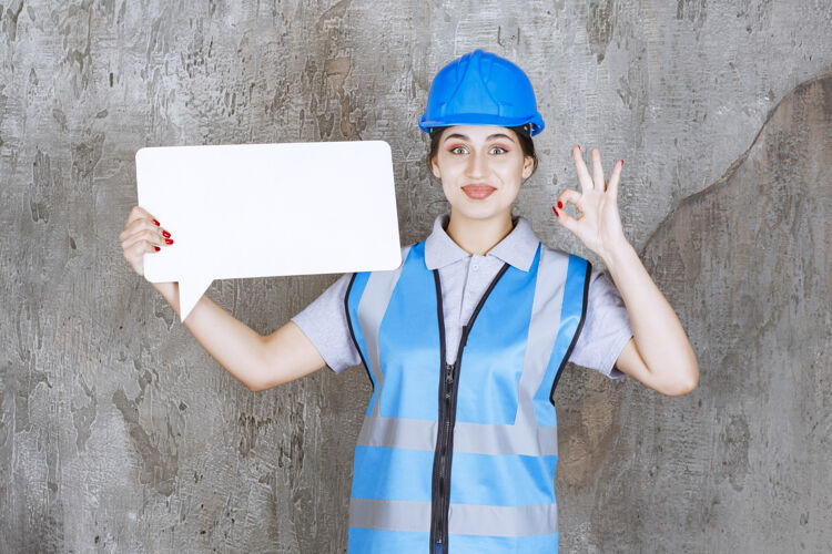安全身穿蓝色制服 头戴安全帽的女工程师 手持一块空白的矩形信息板 并展示享受标志积极交易聪明