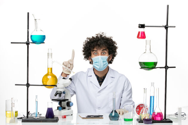 姿势正面图男性化学家穿着医疗服 戴着口罩 坐在白色的桌子上 摆着不同的解决方案溶液化学套装