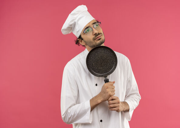 厨师看着一边想着的年轻男厨师 穿着厨师制服 戴着眼镜 拿着煎锅围着下巴男围着壁板