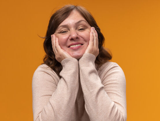 脸颊身穿米色套头衫的中年妇女站在橘色的墙上 两臂贴在脸颊上 面带微笑 快乐而积极高领毛衣站着年龄