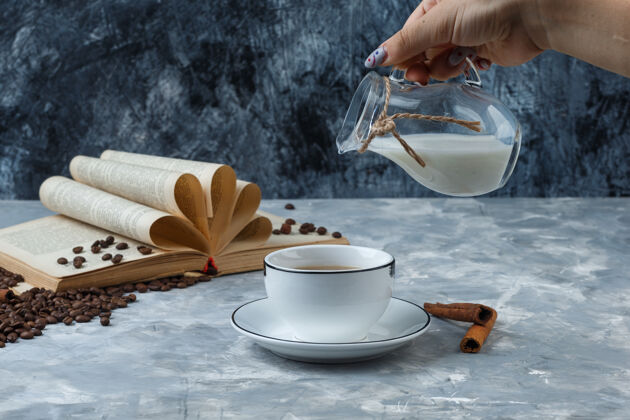 杯子有的用手把牛奶倒进一杯咖啡里 配上咖啡豆 肉桂棒 书上的垃圾和石膏背景 侧视图棍棒自然咖啡