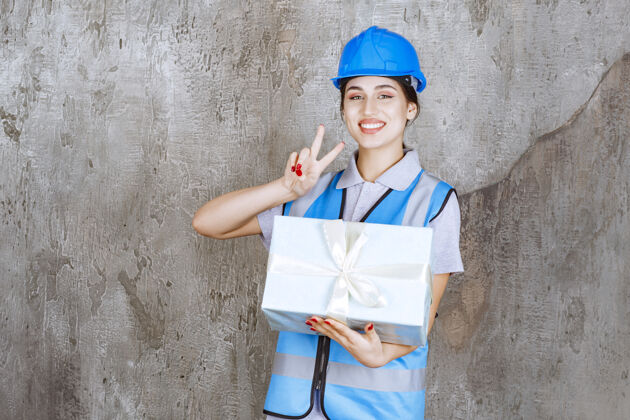 模特身着蓝色制服 头戴安全帽的女工程师 手拿蓝色礼盒 手举正面手势年轻人员工年轻