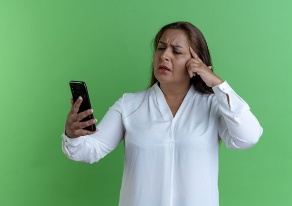 绿色困惑随意的白种中年妇女拿着手机看着手指放在额头上困惑随意额头