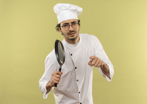 男性自信的年轻男厨师穿着厨师制服 戴着眼镜 拿着煎锅 向你展示手势显示平底锅年轻
