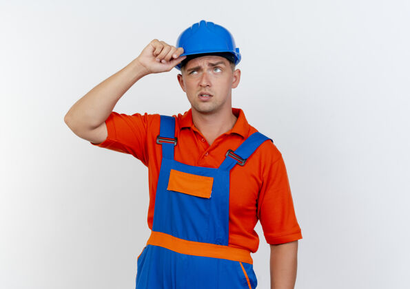 头盔困惑的年轻男性建筑工人穿着制服 戴着安全帽 手放在头盔上戴困惑手