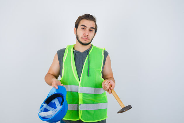 有趣年轻的建筑工人手持头盔和锤子 穿着工作服 看上去很有标志性正视图工装表情制服