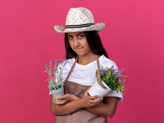 愤怒的穿着围裙 戴着夏帽 手里拿着盆栽的年轻园丁女孩站在粉红色的墙上 脸上露出愤怒的表情围裙年轻的花园