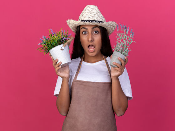 帽子穿着围裙 戴着夏帽 手里拿着盆栽植物的年轻园丁女孩站在粉红色的墙上惊讶不已立场锅年轻的
