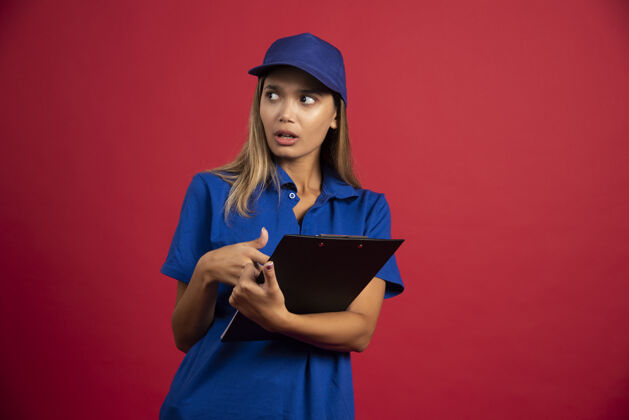 帽子穿着蓝色制服的年轻女子拿着剪贴板和铅笔摆姿势剪贴板指向人