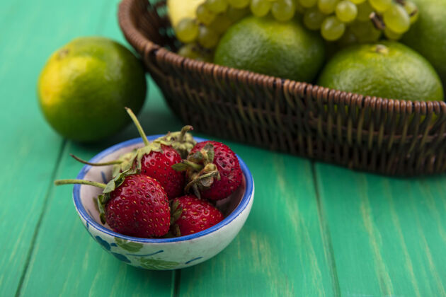 茶碟侧视图草莓在一个茶碟里 橘子和葡萄在一个绿色的墙上的篮子里新鲜橘子食品