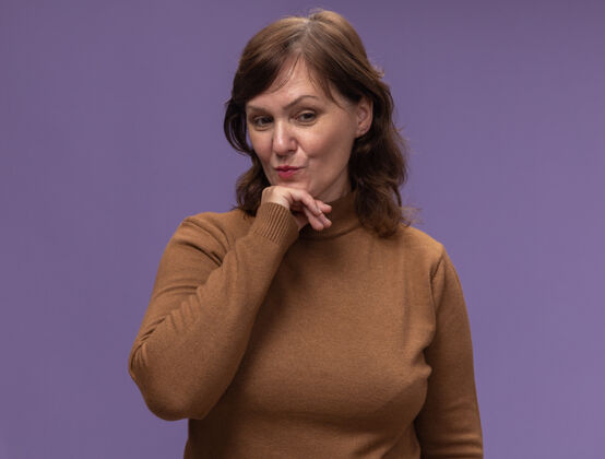 女人穿棕色高领毛衣的中年妇女站在紫色的墙上 手放在下巴上 一边看着一边 一边想着正面年龄思考旁边