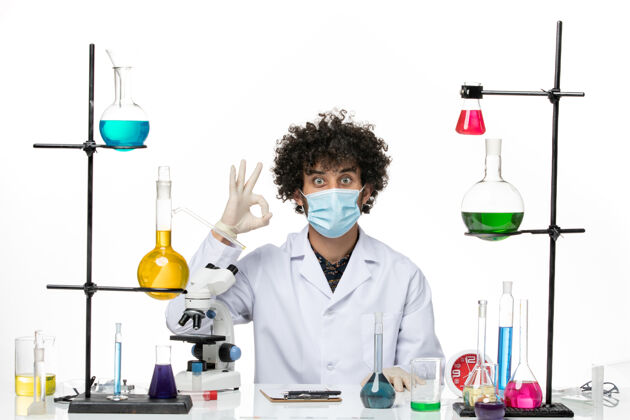 科学正面图男性化学家穿着白色医疗服 戴着口罩 坐在不同的解决方案的白色空间实验室不同化学