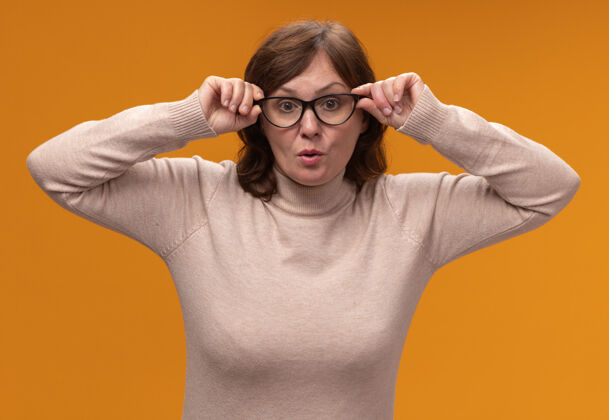 女人穿着米色高领毛衣 戴着眼镜的中年妇女紧紧地站在橙色的墙上站着穿着封闭