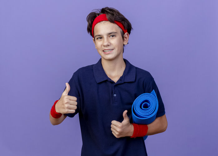 垫子微笑的年轻帅气的运动男孩戴着头带和护腕 戴着牙套 拿着瑜伽垫 竖起大拇指 隔离在紫色的墙上 留着复制空间头带拇指腕带