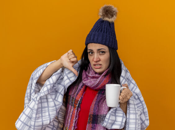 不愉快年轻的白种病女孩戴着冬天的帽子 围着方格围巾 手里拿着一杯茶 拇指朝下 孤零零地站在橙色的墙上拇指显示穿