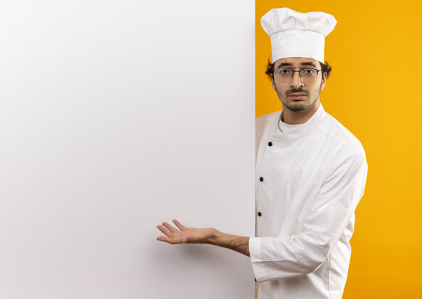 制服年轻的男厨师穿着厨师制服 戴着眼镜 手拿着并展示给隔离在黄色墙壁上的白色墙壁和复印空间不愉快厨师墙