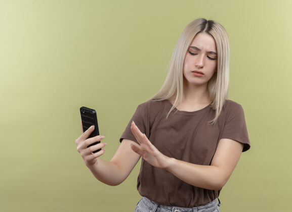 年轻一个年轻的金发女孩拿着手机看着它 在隔离的绿色墙壁上做着“不”的手势不愉快金发手机