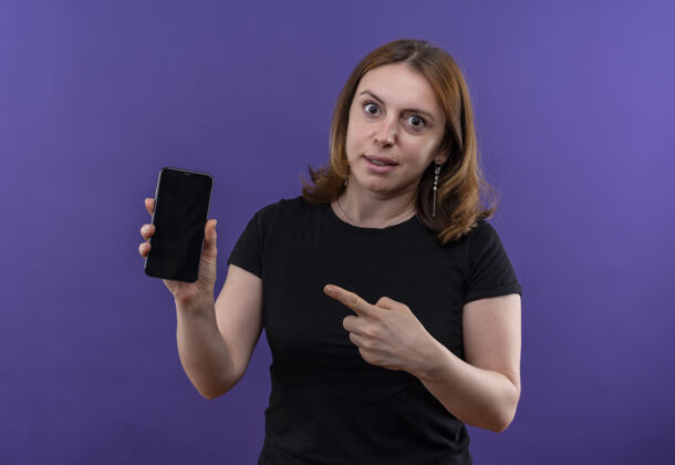 指着惊讶的年轻休闲女子拿着手机 指着孤零零的紫色墙上的手机抱着惊喜女人