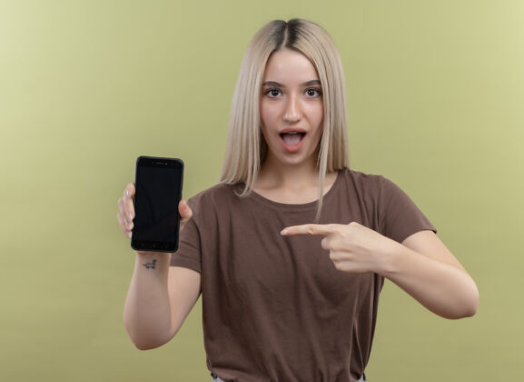 指着一个年轻的金发女孩拿着手机指着孤零零的绿墙上的手机金发惊讶拿着