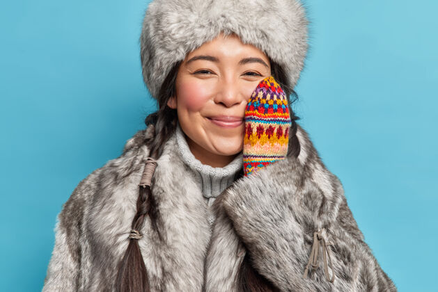 毛衣快乐温柔的爱斯基摩女人穿着冬衣 手放在脸颊上享受冬天的时光 微笑着愉快地对着蓝色的墙壁摆姿势寒冷的气候圣诞节室内东方