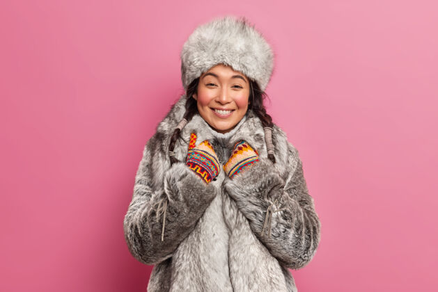 季节穿着灰色皮草外套的漂亮的东方姑娘微笑着真诚地听到有什么愉快的事要在粉色的墙上摆出冬天散步的姿势室内温暖辫子