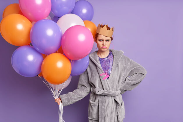 皱眉横拍的是一个穿着柔软睡衣 头戴纸皇冠的不开心女人 手里拿着一堆五颜六色的气球 皱着眉头 脸孤立地贴在紫色的墙上脸衣服漂亮