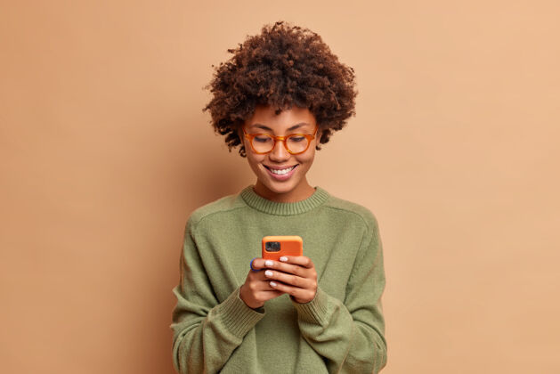 人《女人的孤立镜头》使用智能手机应用程序享受浏览社交媒体制作新闻内容制作在线订单戴眼镜和休闲套头衫在米色工作室墙上摆姿势肖像眼镜青少年