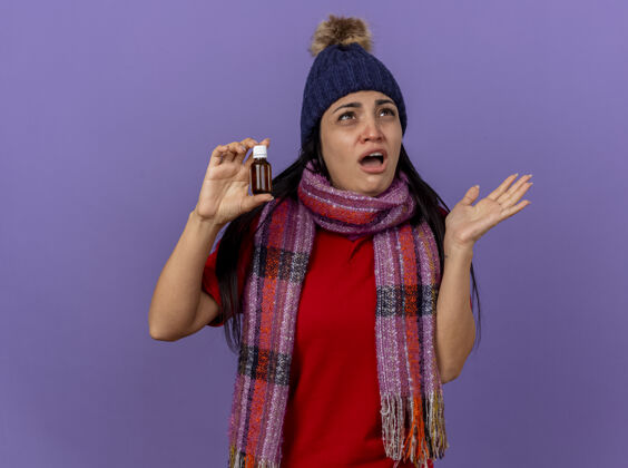 年轻令人印象深刻的年轻白种女孩戴着冬天的帽子和围巾拿着药剂在玻璃上抬头看显示空手孤立在紫色的墙壁与复制空间显示药物围巾