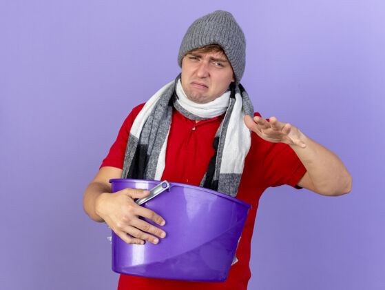 年轻年轻英俊的金发病男子戴着冬天的帽子和围巾 手里拿着塑料桶 手伸向空中 隔离在紫色的墙上 留着复制空间疾病表达围巾