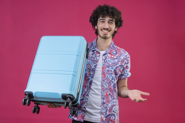 手微笑着的年轻英俊的卷发旅行者男人拿着手提箱 在孤立的粉色墙上展示着空手年轻秀帅气